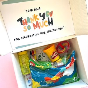 May include: A white box with a thank you card inside. The card says "Dear Aria, Thank You So Much for celebrating our special day!" The box is filled with colorful toys, including a container of Play-Doh, a spinning top, crayons, and a bubble bottle.