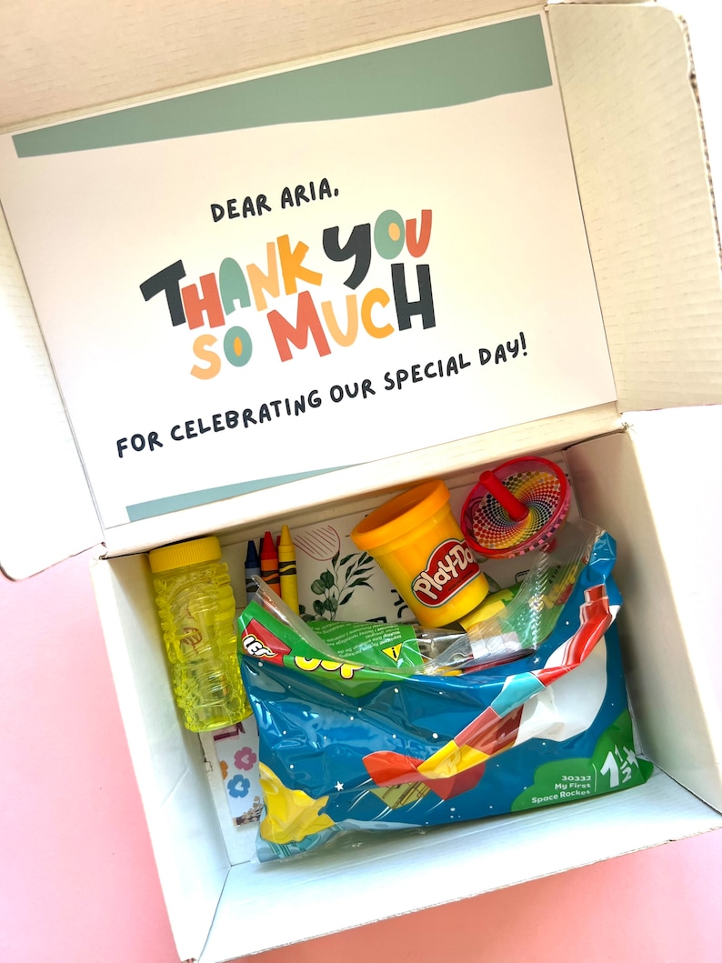 May include: A white box with a thank you card inside. The card says "Dear Aria, Thank You So Much for celebrating our special day!" The box is filled with colorful toys, including a container of Play-Doh, a spinning top, crayons, and a bubble bottle.