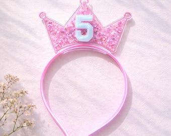 May include: A pink crown-shaped headband with the number "5" in light blue. The crown is filled with pink glitter and has a matching pink headband. The image is taken on a soft, light pink surface.