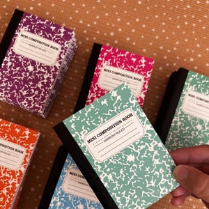 May include: A selection of miniature composition notebooks in shades of purple, pink, green, orange, and blue. Each notebook has a black spine and a white label with the text "MINI COMPOSITION BOOK - NARROW RULED". The notebooks feature a classic speckled pattern.