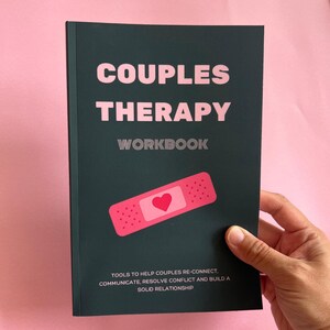 May include: A dark green couples therapy workbook with a pink plaster with a heart on it. The text on the cover reads "COUPLES THERAPY WORKBOOK" and "TOOLS TO HELP COUPLES RE-CONNECT, COMMUNICATE, RESOLVE CONFLICT AND BUILD A SOLID RELATIONSHIP."