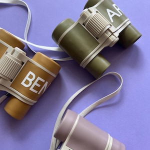 May include: Three pairs of toy binoculars with a strap. The binoculars are in green, brown, and purple. The binoculars have the names "AR", "BEN", and "MIA" written on them.