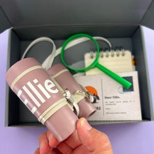 May include: A pink and white children's binoculars with the name "Ellie" printed on the side. The binoculars are in a gray box with a green magnifying glass, a notebook, and a postcard.