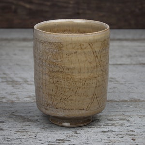 May include: A ceramic mug with a light brown glaze and a map design. The mug has a wide base and a rounded shape.