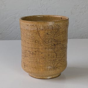 May include: A tan ceramic tumbler with a map design. The cylindrical cup has a slightly flared rim and a rounded base. The map design is etched into the surface, with continents and lines visible. The cup is approximately 4 inches tall.