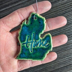 May include: A green and blue ceramic ornament shaped like the state of Michigan with the word "Home" written in black script.