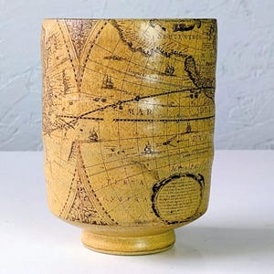 May include: A cylindrical, beige ceramic cup with a map design. The map is in brown and features nautical charts and geographical details. The cup has a small, round base and a slightly textured surface.