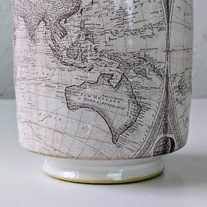 May include: A ceramic mug with a vintage world map design. The off-white mug features a detailed map in shades of brown and gray, with a white base and a yellow rim. The mug has a classic, slightly flared shape.