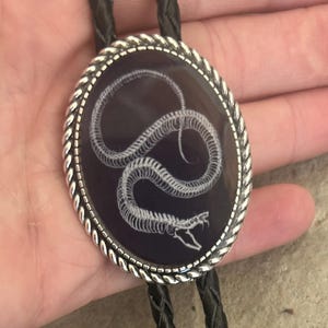 Könnte beinhalten: Ovales Bolo-Krawatte mit weißem Schlangenmuster auf schwarzem Hintergrund. Der silberfarbene Rahmen hat einen seilartigen Rand. Das schwarze geflochtene Band hat dekorative silberfarbene Spitzen. Ein einzigartiges Accessoire.