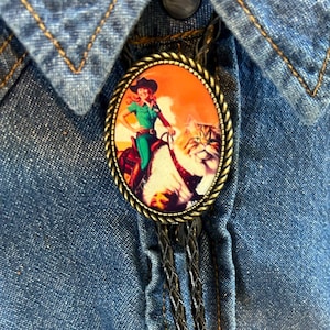 Puede incluir: Un corbatín bolo con un cordón de cuero marrón y un colgante ovalado de metal con una ilustración vintage de una mujer montando un tigre. El colgante tiene un marco dorado.