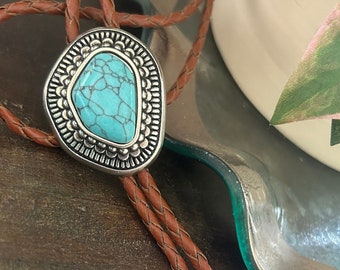 May include: A brown leather bolo tie with a silver and turquoise stone pendant. The pendant is a teardrop shape with a detailed design.