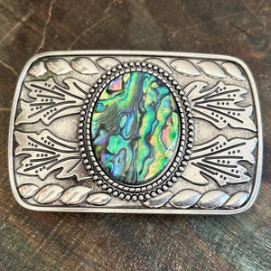 Puede incluir: Hebilla de cinturón rectangular plateada con un centro ovalado de concha de abulón. La concha muestra tonos iridiscentes verdes, azules y amarillos. La hebilla tiene un patrón decorativo de hojas y puntos en los bordes.