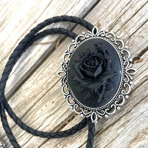Può includere: Un bolo tie nero con un pendente ovale argentato che presenta una rosa nera con dettagli intricati. Il pendente ha un design a filigrana decorativo.