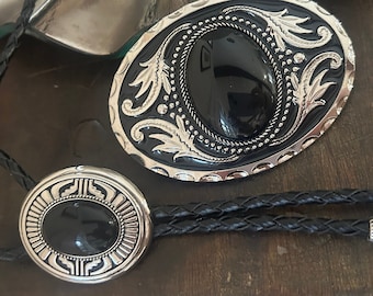 May include: An oval-shaped, silver-toned belt buckle and a bolo tie with black braided cord. The buckle features a black oval center stone, surrounded by a silver filigree design. The bolo tie has a matching black stone and silver accents.