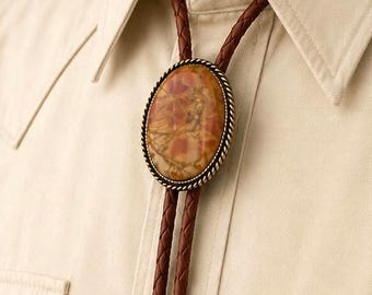 Puede incluir: Una corbata bolo con una piedra central ovalada de patrones jaspeados en marrón y beige, enmarcada en metal trenzado. La corbata consta de un cordón de cuero trenzado marrón terminado con dos puntas metálicas decorativas a juego.