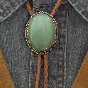 May include: A bolo tie featuring a large, oval, pale green stone in a decorative metal frame. It has braided brown leather cords and ornate metal tips, set against a denim shirt and brown suede vest.