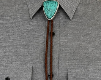 May include: A bolo tie featuring a triangular turquoise stone with black veining set in a silver frame. The pendant is attached to a braided brown leather cord that ends in two silver-toned metal tips, worn over a grey button-down shirt.