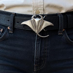 May include: A black leather belt with a textured surface and a silver-toned metal buckle shaped like a manta ray. The belt is worn through the loops of dark blue denim jeans, paired with a light-colored button-down shirt with visible buttons.
