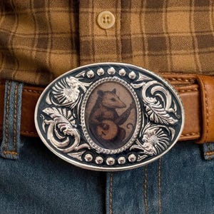 Pode incluir: Fivela de cinto ovalada prateada com cinto de couro castanho. A fivela apresenta uma imagem central de um gambá tocando banjo, rodeada por ornamentos prateados. Usada com calças de ganga azuis e camisa xadrez castanha.