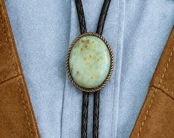 May include: A bolo tie featuring a smooth, oval-shaped light green stone with brown speckles, set in a textured bronze-toned metal frame. The pendant is attached to a black braided leather cord, worn over a light blue button-down shirt and brown vest.