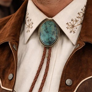 May include: A close-up of a bolo tie with a turquoise stone pendant, brown braided leather cord, and ornate metal tips. The tie is set against a cream-colored shirt and a brown suede jacket with button details. The shirt collar has embroidered details.