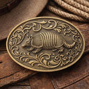 Puede incluir: Hebilla de cinturón ovalada de latón antiguo con un diseño detallado de armadillo y adornos ornamentales. La hebilla descansa sobre un cinturón de cuero marrón, con un sombrero de vaquero y una cuerda en el fondo. Accesorio de estilo occidental.
