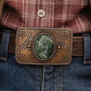 Puede incluir: Primer plano de una hebilla de cinturón con una piedra verde ovalada y detalles florales. La hebilla se lleva con un cinturón de cuero marrón, vaqueros azules y una camisa a cuadros roja.