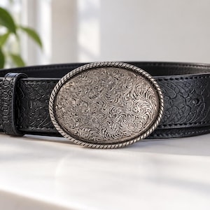 Puede incluir: Un cinturón negro con un patrón texturizado y en relieve en la correa y una gran hebilla ovalada de metal plateado. La hebilla está decorada con intrincados grabados florales y está enmarcada por un borde con diseño de cuerda trenzada.