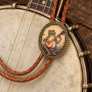 May include: A bolo tie featuring an oval-shaped pendant with a rope-style brass border. The pendant displays an illustration of a green frog playing a stringed instrument. The tie consists of two braided brown leather cords attached to the pendant.