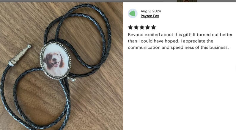 Puede incluir: Un corbat&iacute;n de cuero negro con un colgante ovalado de plata que presenta una foto de un perro marr&oacute;n y blanco. El colgante tiene un borde de plata.