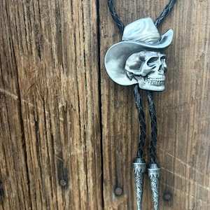 May include: A bolo tie featuring a silver-toned metal skull wearing a cowboy hat as the central slide. The cord is made of black braided leather and terminates in two matching silver-toned metal tips with intricate engraved floral patterns.