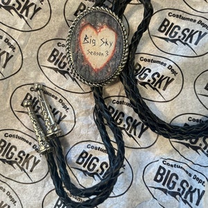 Puede incluir: Un corbat&iacute;n bolo negro con un colgante de plata en forma de coraz&oacute;n. El colgante tiene un dise&ntilde;o de veta de madera y el texto "Big Sky Season 3" escrito en &eacute;l.