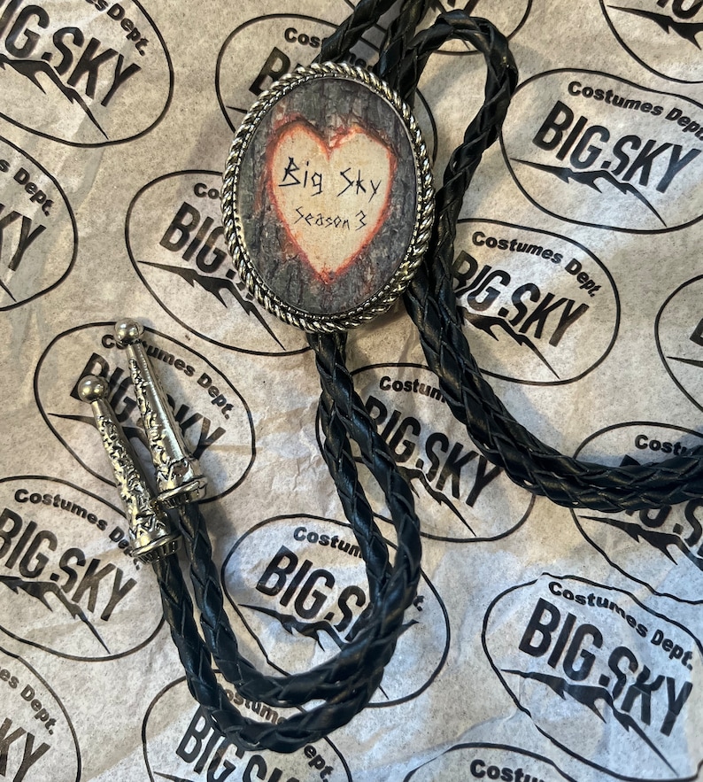Puede incluir: Un corbat&iacute;n bolo negro con un colgante de plata en forma de coraz&oacute;n. El colgante tiene un dise&ntilde;o de veta de madera y el texto "Big Sky Season 3" escrito en &eacute;l.