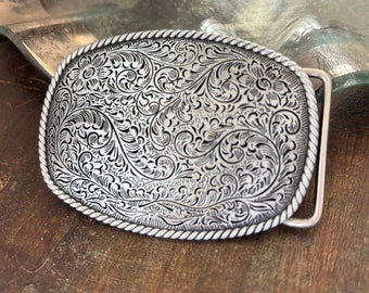May include: Silver-toned belt buckle with an intricate floral design. The buckle has a rope-like border.