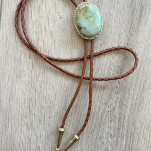 Pode incluir: Uma gravata bolo com uma pedra oval verde claro com uma borda dourada em forma de corda. O cord&atilde;o de couro tran&ccedil;ado castanho tem pontas de metal dourado. A gravata &eacute; exibida em uma superf&iacute;cie de madeira clara.