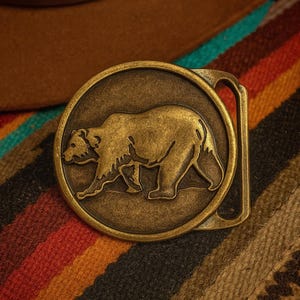 Puede incluir: Hebilla de cinturón redonda de latón antiguo con un diseño detallado de oso. La hebilla se encuentra sobre un fondo de sombrero marrón y un textil a rayas de colores, mostrando una estética rústica.