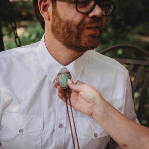 Pode incluir: Uma camisa branca com uma gravata bolo turquesa. A gravata bolo tem uma borda prateada em volta da pedra turquesa. O cord&atilde;o da gravata &eacute; castanho.
