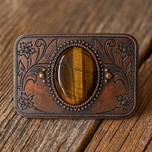 May include: A rectangular, copper-toned belt buckle with an oval tiger's eye stone at the center. The buckle features floral and scrollwork designs, with a rope-like border around the stone and the outer edge.