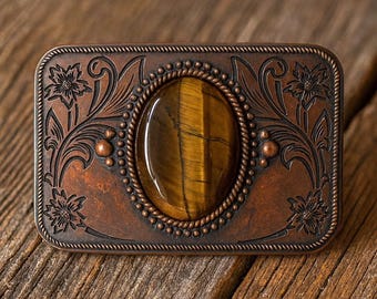 Puede incluir: Hebilla de cinturón rectangular de color cobre con una piedra de ojo de tigre ovalada en el centro. La hebilla presenta diseños florales y de volutas, con un borde en forma de cuerda alrededor de la piedra y el borde exterior.