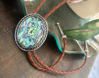 May include: A brown leather bolo tie with a silver oval pendant featuring a green, blue, and iridescent abalone shell.
