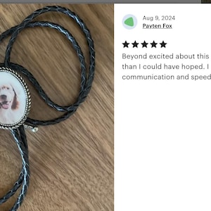 Puede incluir: Un corbat&iacute;n de cuero negro con un colgante ovalado de plata que presenta una foto de un perro marr&oacute;n y blanco. El colgante tiene un borde de plata.