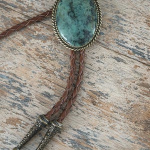 May include: A bolo tie featuring a large, oval turquoise stone set in a decorative brass-colored frame. The braided leather cord is brown, with decorative metal tips. The bolo tie rests on a weathered wooden surface.