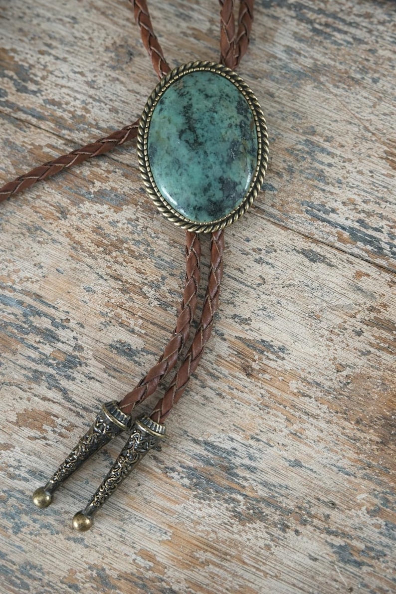 May include: A bolo tie featuring a large, oval turquoise stone set in a decorative brass-colored frame. The braided leather cord is brown, with decorative metal tips. The bolo tie rests on a weathered wooden surface.