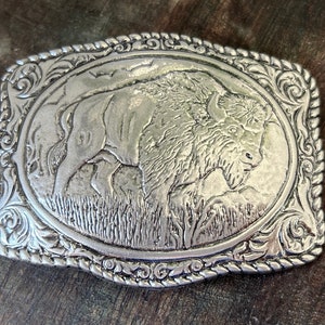 Puede incluir: Hebilla de cinturón de plata con un diseño ornamentado que presenta una imagen detallada de un búfalo en un campo de hierba.