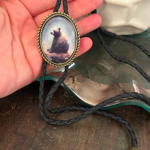 May include: A black bolo tie with a round, bronze-colored pendant featuring a detailed image of a brown bear. The pendant has a textured, rope-like border.