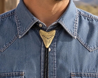 Op de afbeelding: Een bolo-das met een metalen goudkleurige haaientandhanger in het midden van een zwart gevlochten leren koord. De hanger heeft een getextureerd oppervlak met zichtbare ribbels en groeven, gedragen op een blauw spijkerhemd met drukknopen.