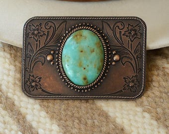 May include: A rectangular copper-colored belt buckle with an oval turquoise stone center. The buckle features floral and scrollwork designs around the stone and edges. The background includes a cream-colored hat and a patterned fabric.