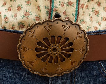 May include: A brown leather belt with a decorative bronze buckle, featuring a floral design with triangular details. The belt is worn with blue denim jeans and a floral shirt. The buckle's diameter is approximately 10 cm.