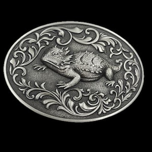 May include: Silver oval belt buckle with a detailed relief of a horned lizard. The lizard is surrounded by ornate, swirling floral patterns. The buckle has a vintage, antique appearance, with a dark background.