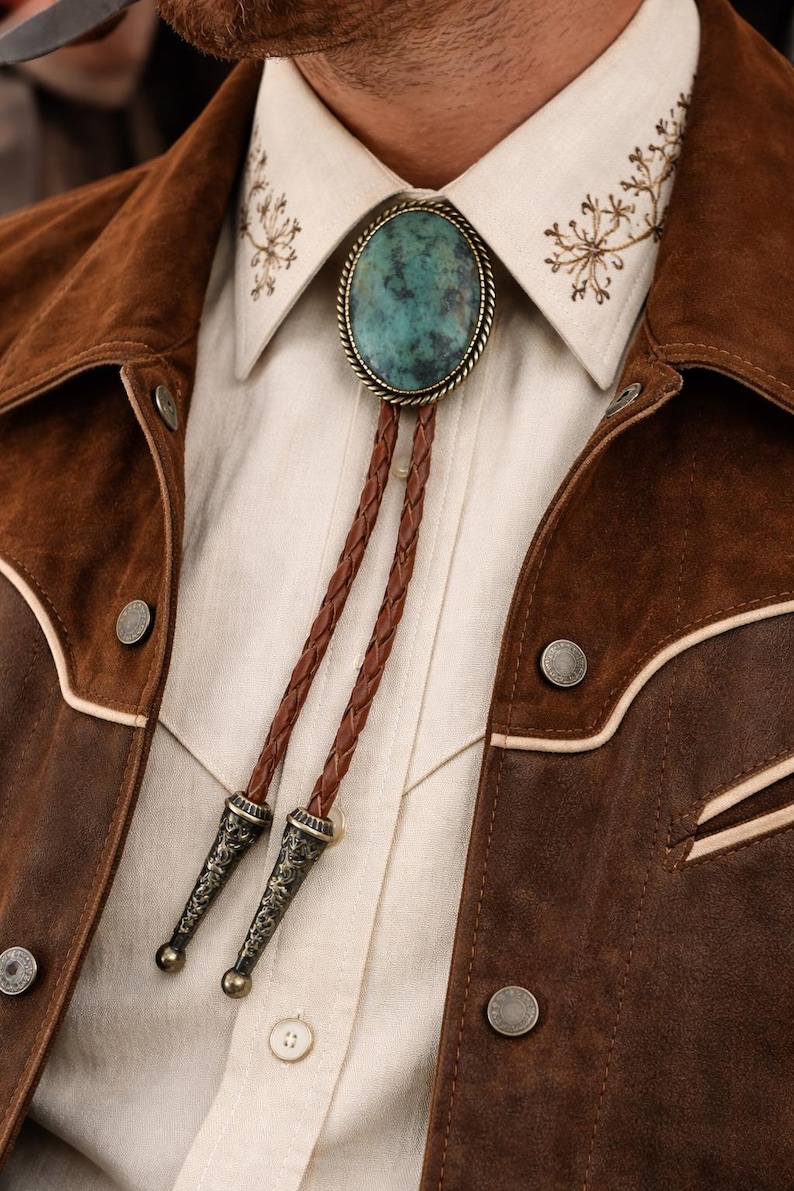 May include: A close-up of a bolo tie with a turquoise stone pendant, brown braided leather cord, and ornate metal tips. The tie is set against a cream-colored shirt and a brown suede jacket with button details. The shirt collar has embroidered details.